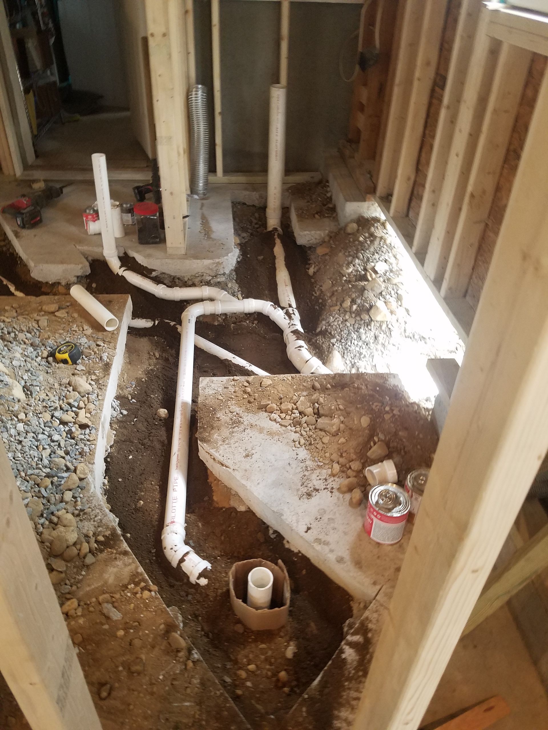 Exposed plumbing and pipes in a house under construction, with dirt floor and a concrete shower base.