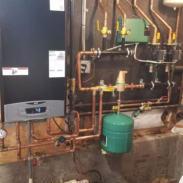 Boiler room with copper pipes, valves, and a wall-mounted heating unit above a green tank.