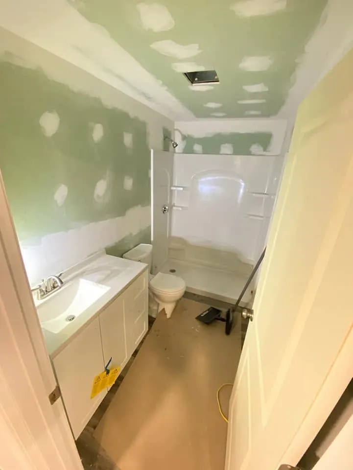 Narrow bathroom with green drywall, white sink vanity, toilet, bathtub, and unfinished ceiling patching.