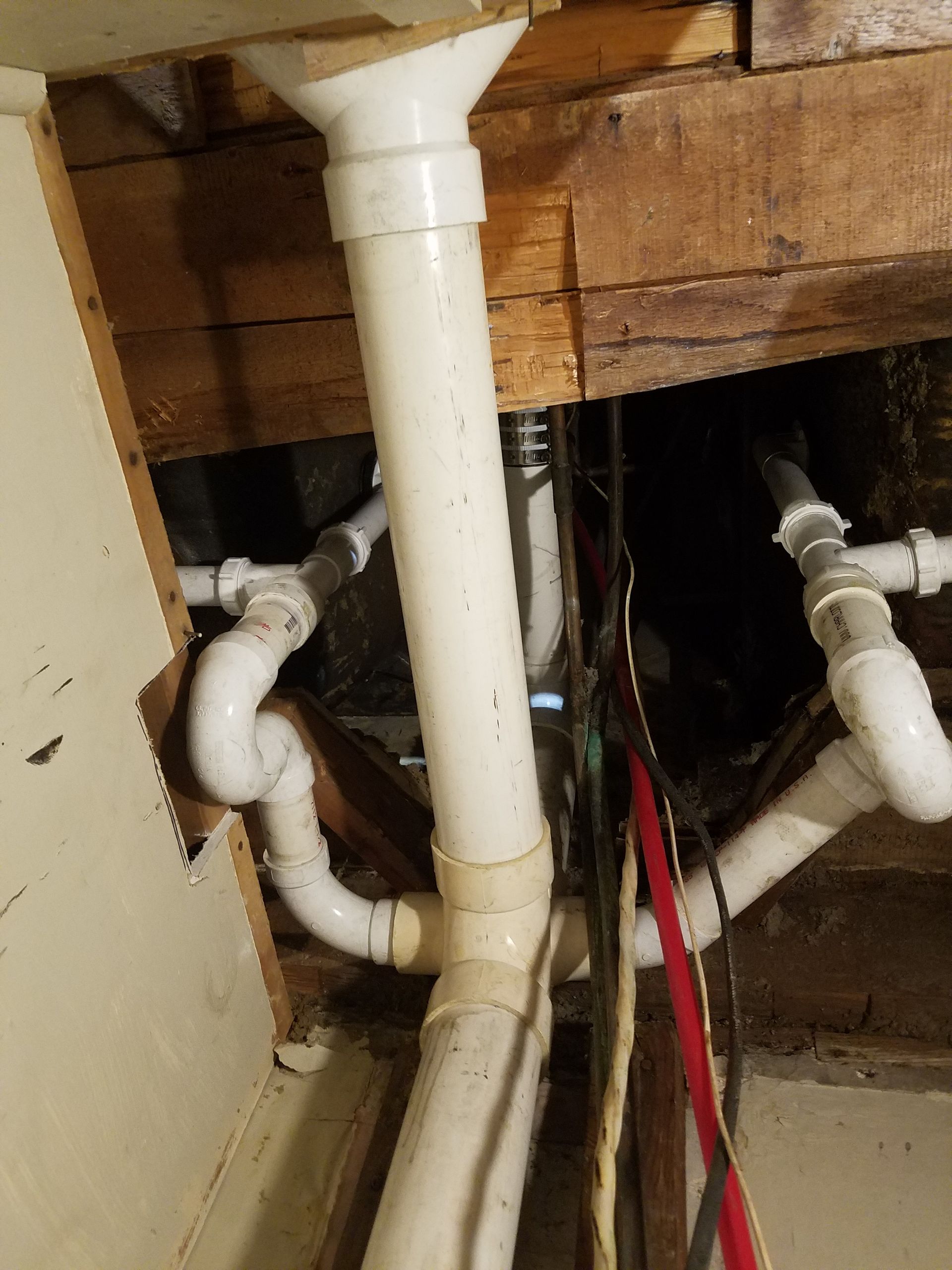 Exposed white plumbing pipes and fittings beneath a sink or floor opening, with wooden joists above.