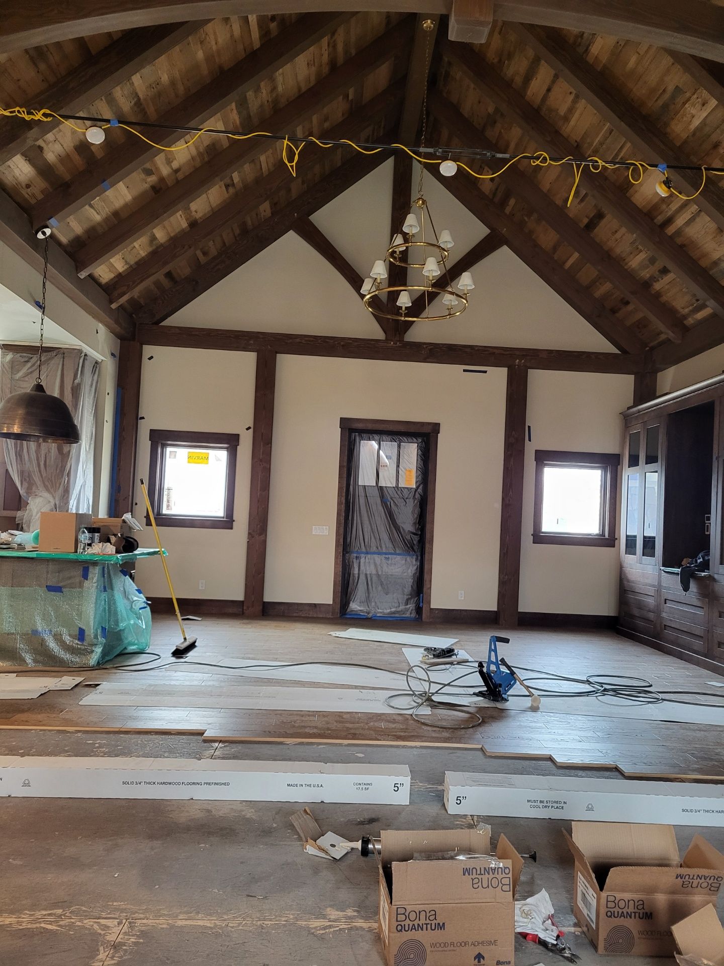 A room under construction with a vaulted ceiling, exposed wooden beams, a central chandelier, and scattered work supplies.