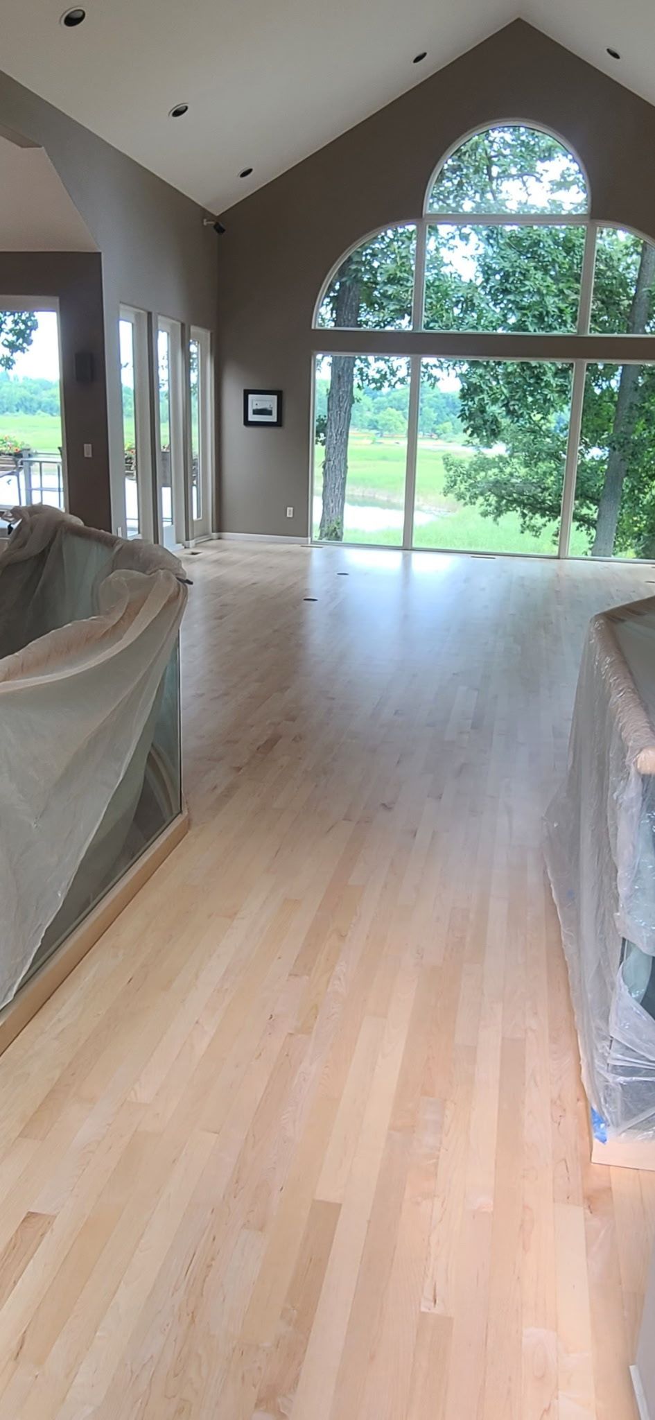 An open, sunlit room with light wood floors and vaulted ceilings, overlooking trees through a large arched window.