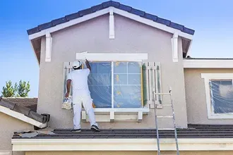 Person painting window trim on a two-story house, using a ladder. The house has a beige exterior and a blue roof.
