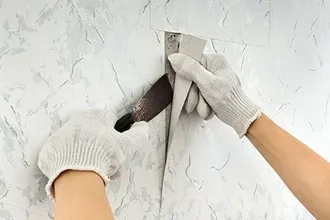 Person wearing gloves cutting wallpaper with a blade along a wall seam.