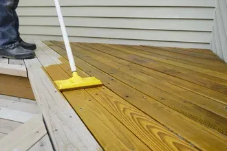 Person staining a wooden deck with a yellow applicator. Light brown stain applied to the wood.
