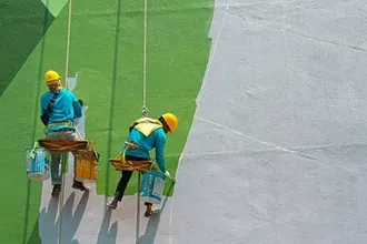 Two painters in safety gear, hanging and painting a large wall green and white.