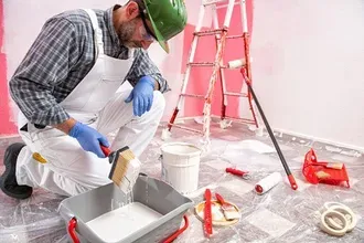 Painter kneels, dipping a brush into paint tray, preparing to paint a room; ladder and paint cans nearby.