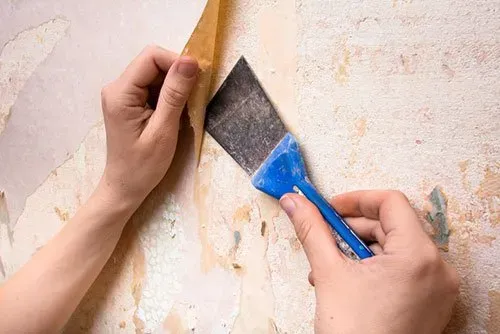 Hands peeling wallpaper with a scraper tool.