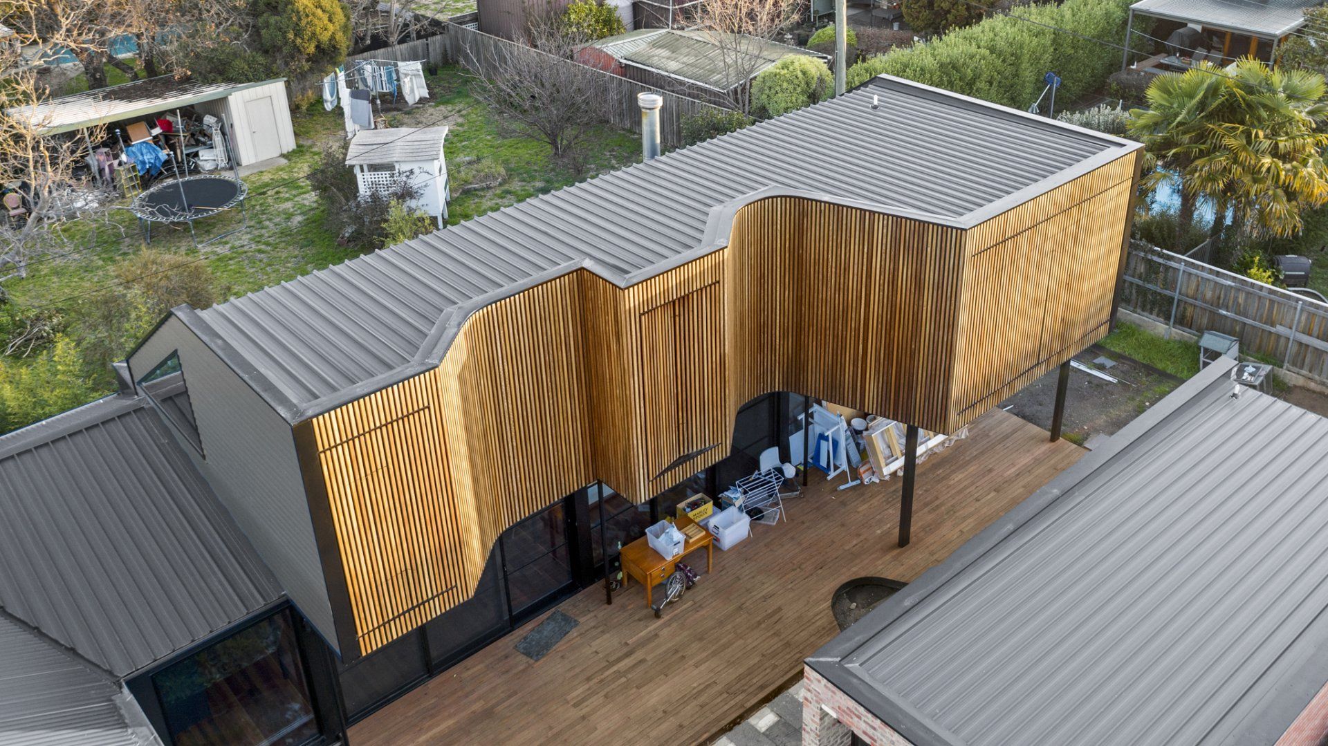 Modern house with curved, wood-paneled facade; gray metal roof; wooden deck; yard with trampoline.