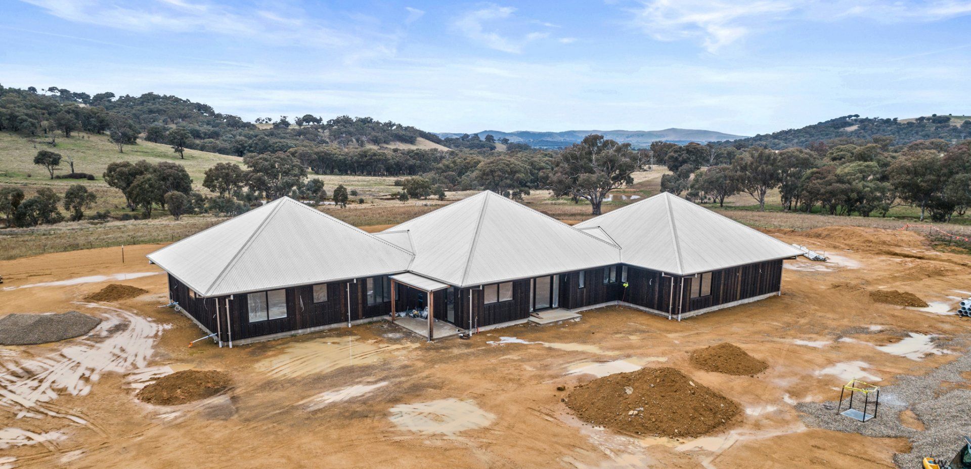 A house under construction on a dirt lot, with a surrounding landscape of hills and trees.