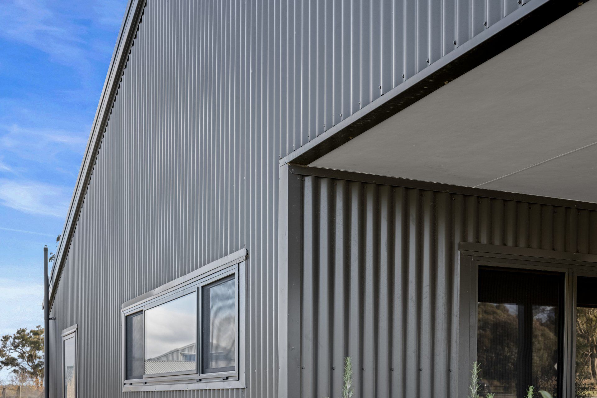 Silver corrugated metal building with white-framed window and overhang against a blue sky.
