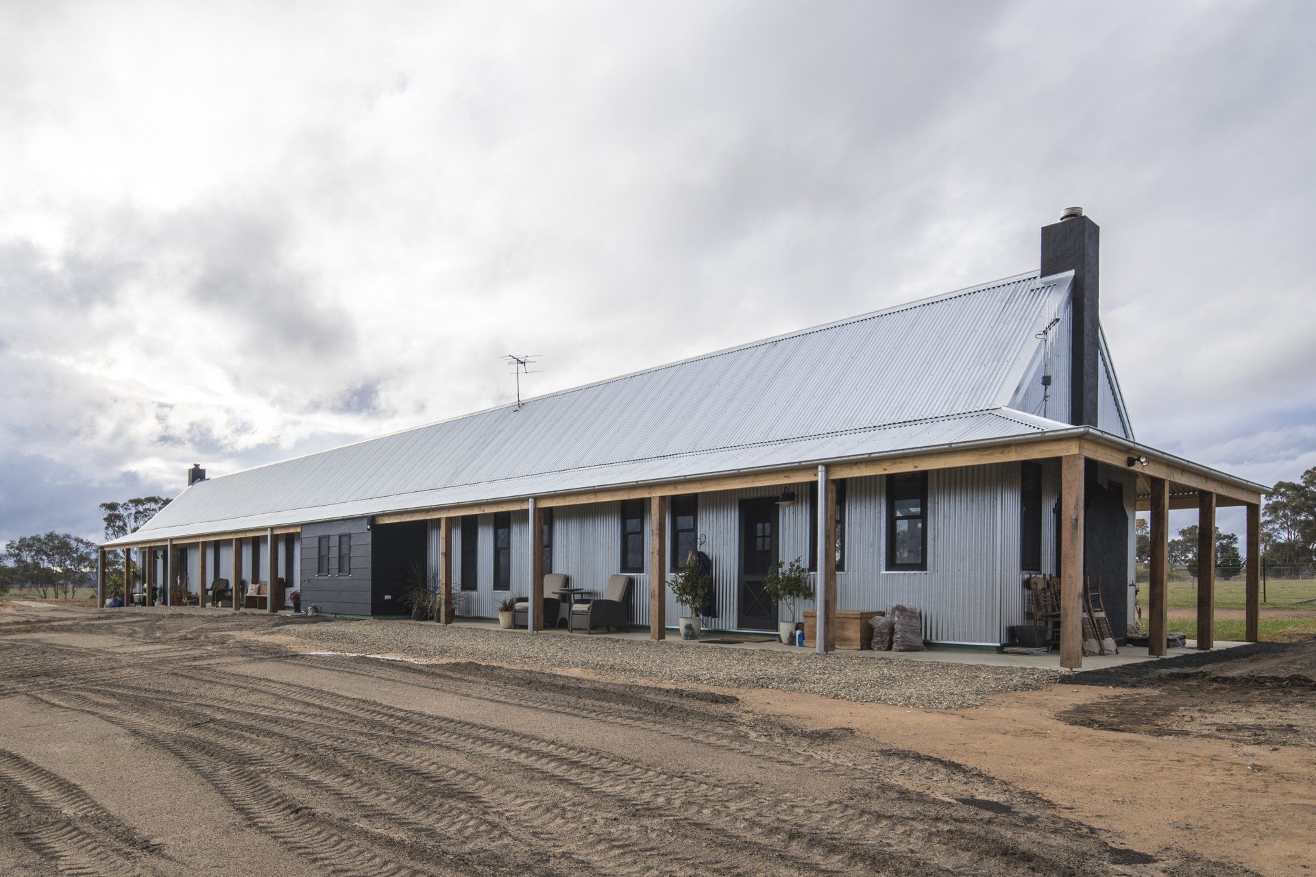 Long, low building with a corrugated metal roof and a porch. The building is gray with a dirt driveway in front.