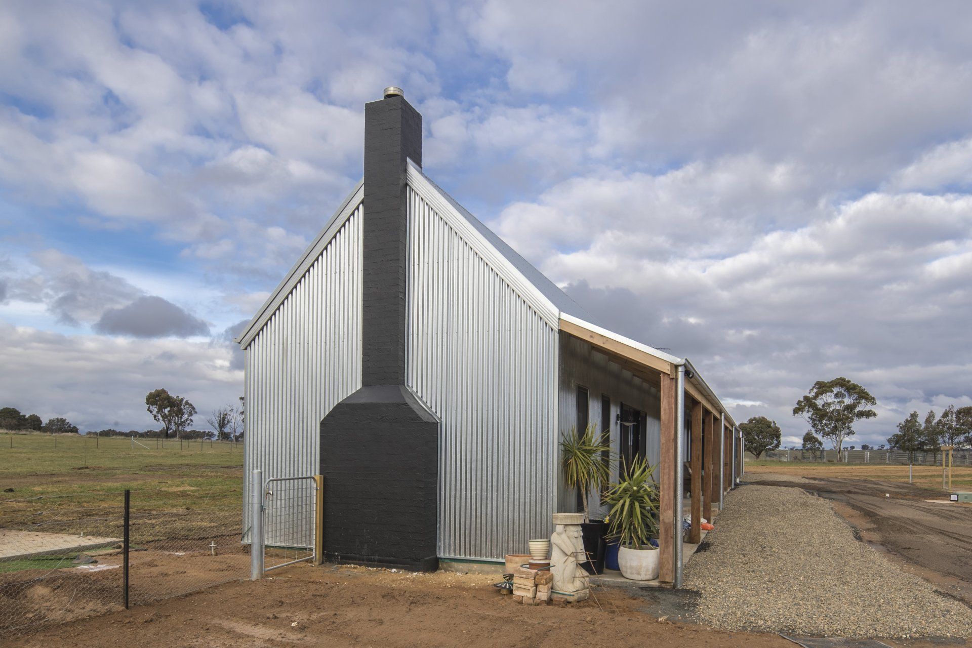 A modern, metal-clad home with black chimney, long porch, set in a rural landscape under a cloudy sky.