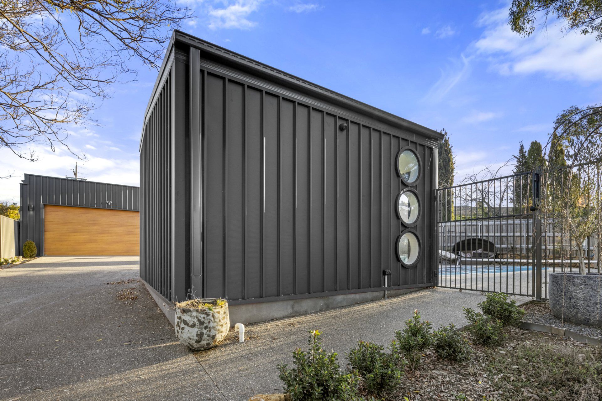Modern, dark grey building with circular windows, gravel driveway, and gate to a pool.
