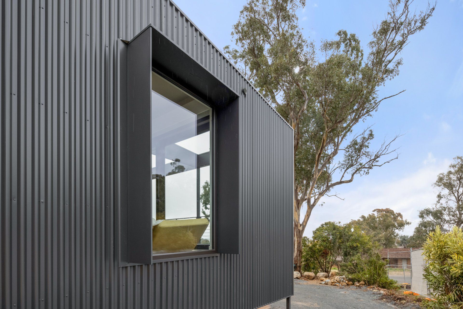 Dark gray corrugated metal exterior of a modern building with a large window and black frame, set against a tree and blue sky.