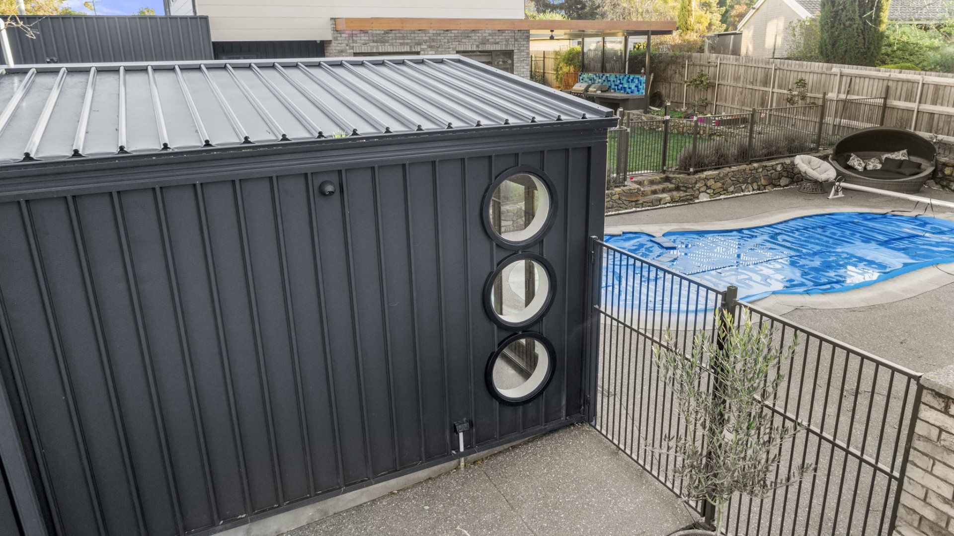 Dark gray shed with circular windows, metal roof, beside a pool covered in blue tarp.