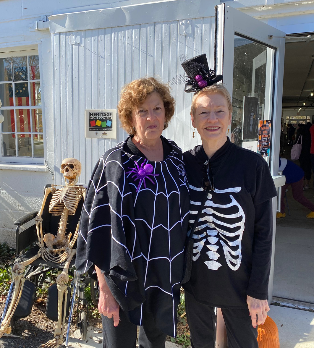 Two women in Halloween costumes pose outside a store. One wears a spider web poncho, the other a skeleton suit.