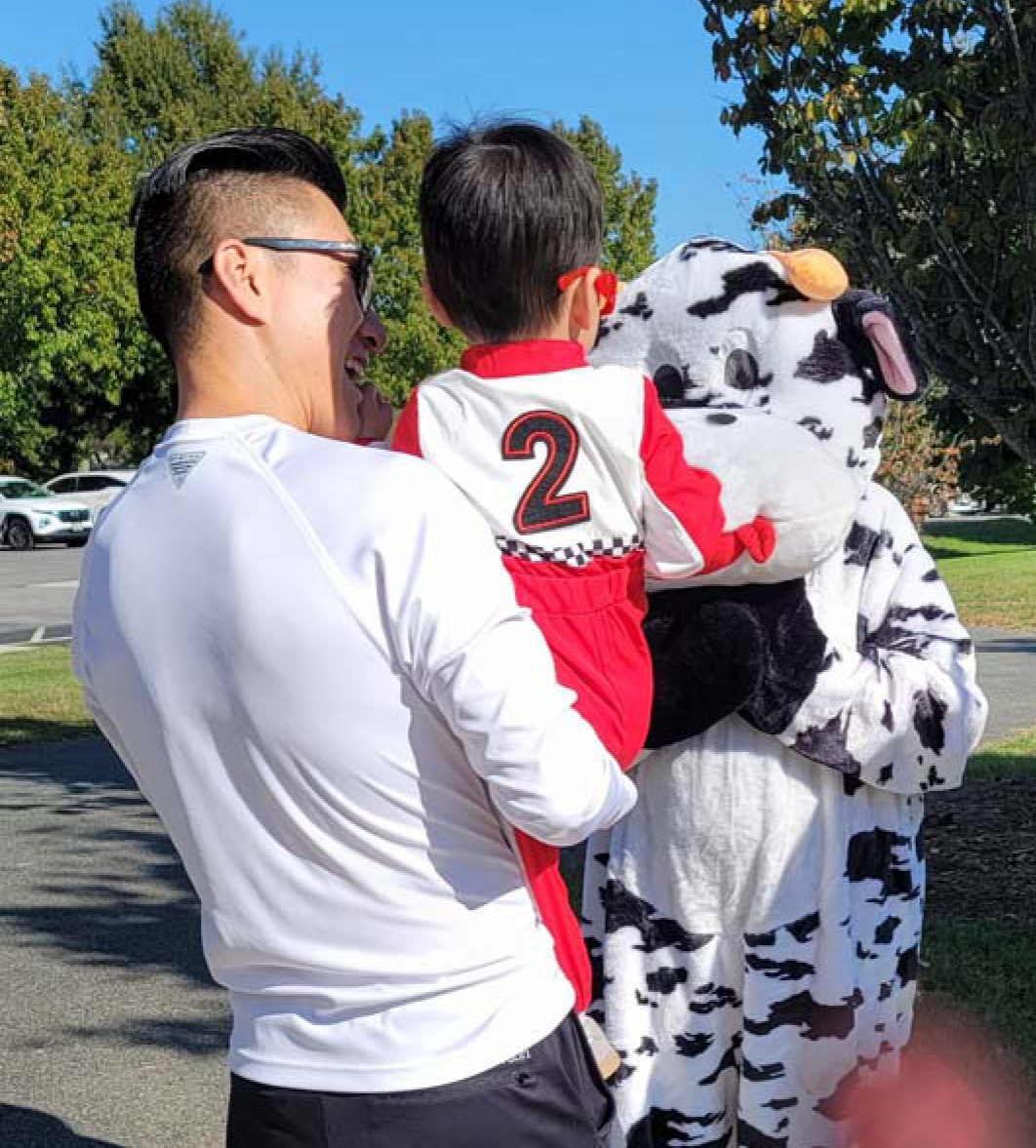 Man holding a child in a racing suit, with a Cow mascot. Outdoors on a sunny day.