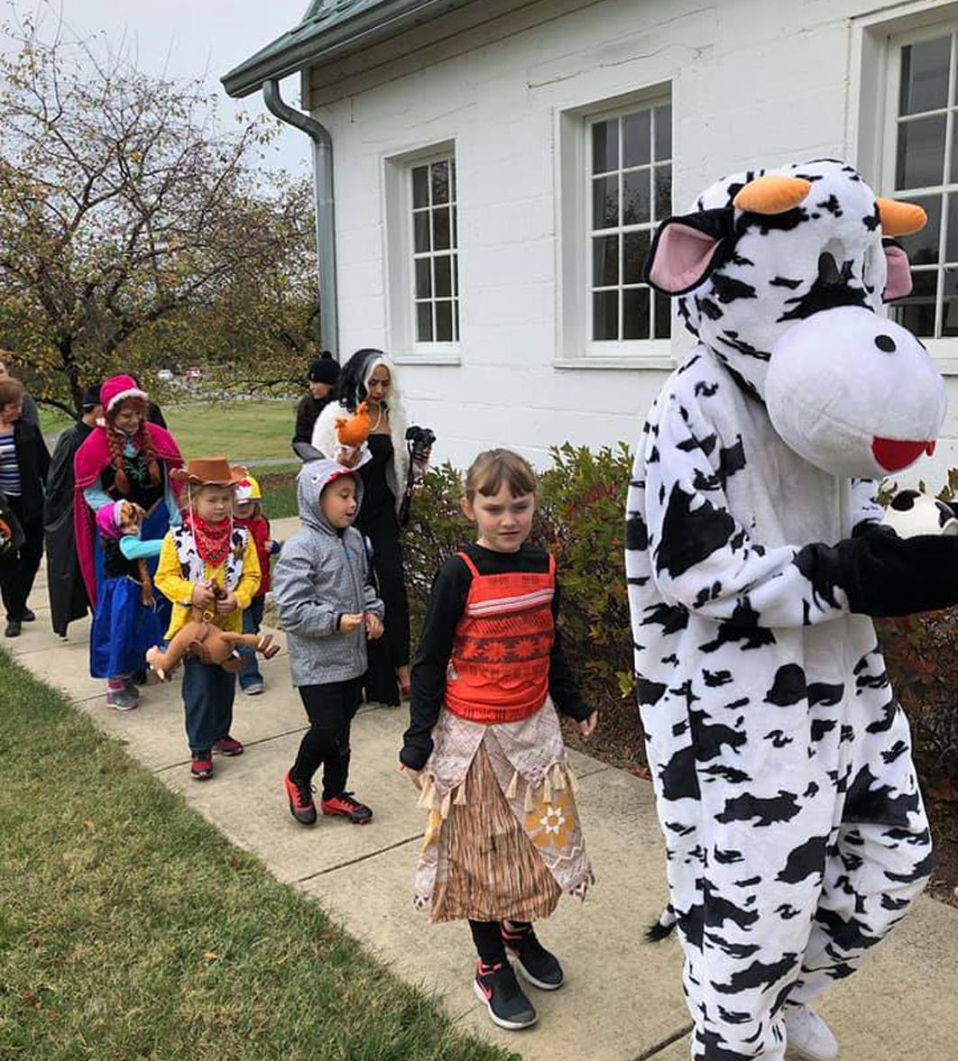Children in costumes parade outdoors next to a cow mascot, following a sidewalk along a building.