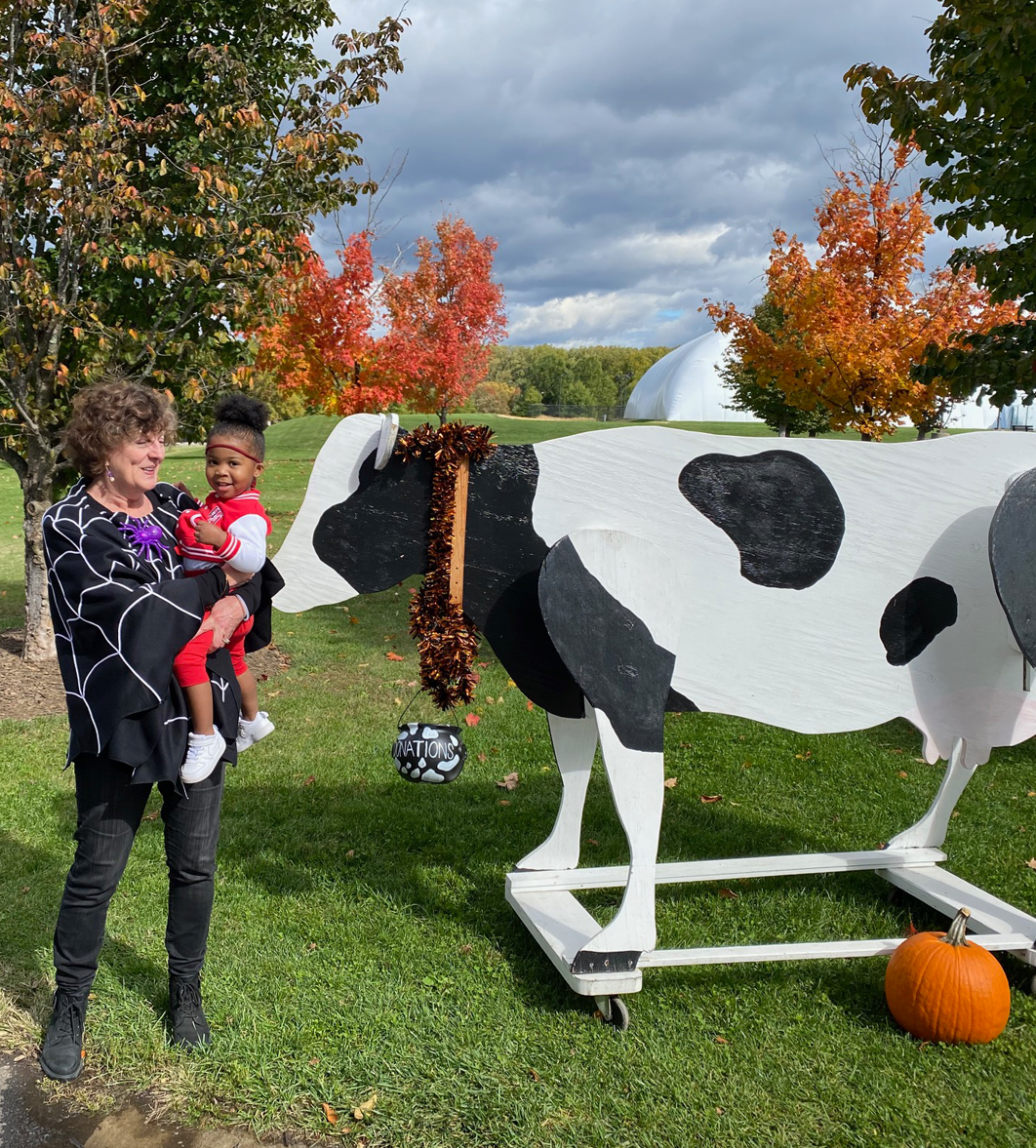 Woman in spiderweb shawl holding a child near a large cow statue and pumpkin. Autumn leaves in the background.