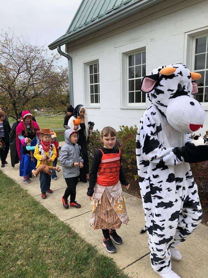 A group of children in costumes are walking down a sidewalk next to a cow mascot.