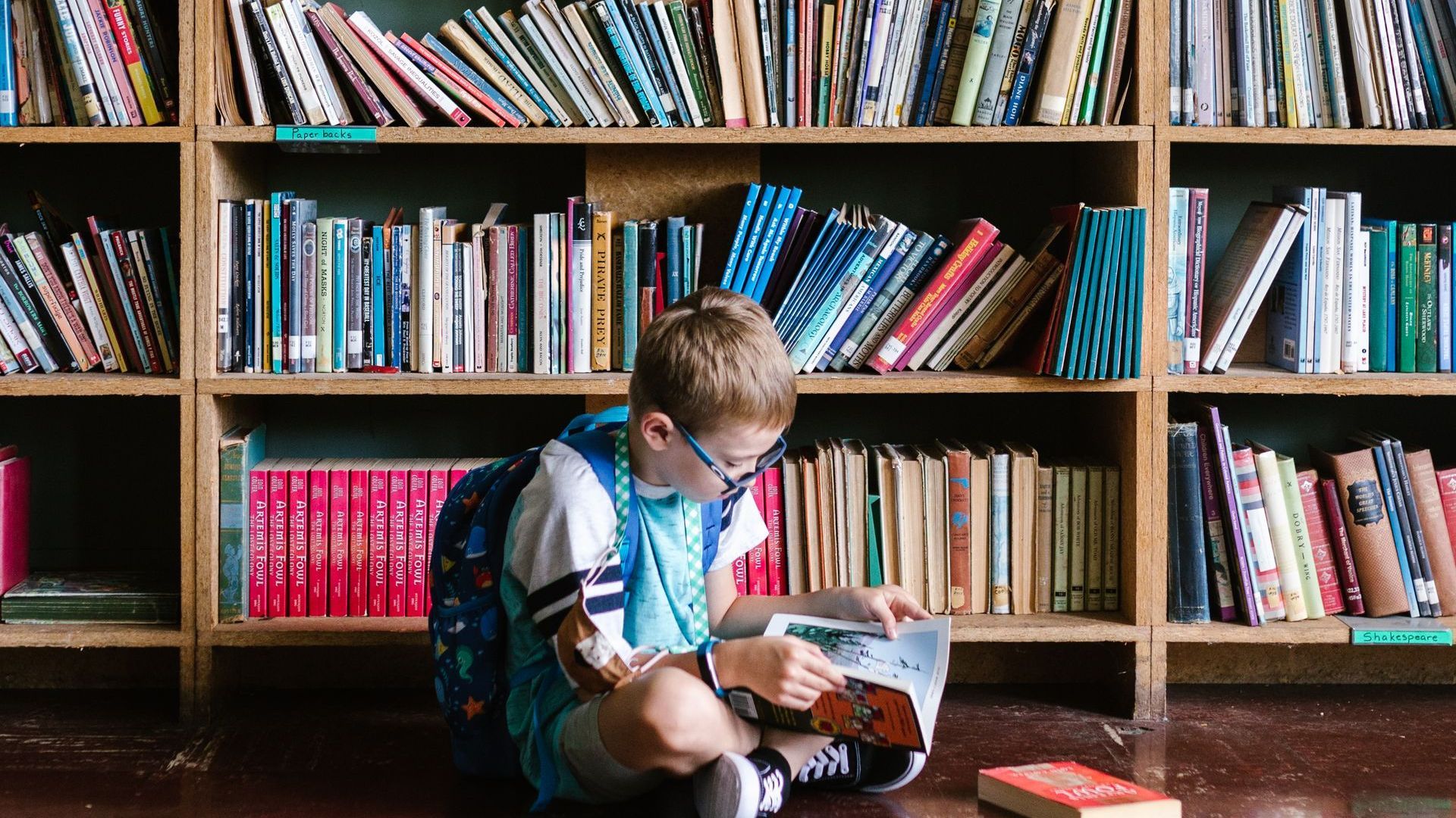 A young boy is sitting on the floor reading a book in the MOOseum library.