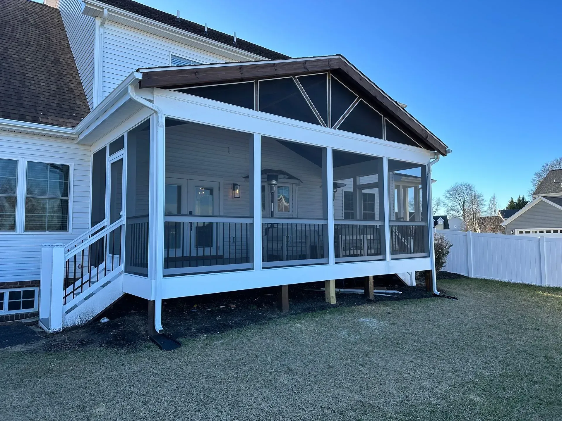 White screened porch with railing attached to a house, seen from the yard on a sunny day.
