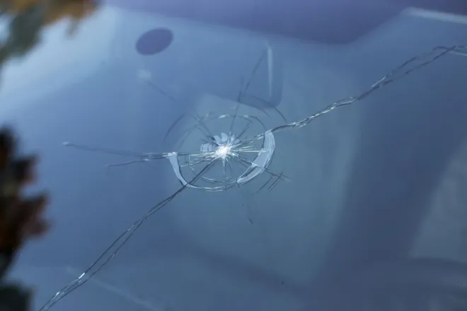 La mano di una persona stringe il volante davanti al parabrezza di un'auto, che presenta una grande crepa a forma di stella causata dall'impatto con un sasso.