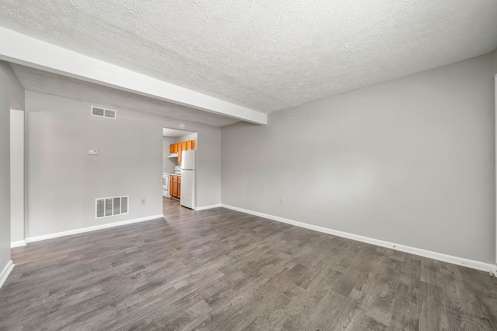 Empty gray living room with carpeted floor, white ceiling beam, and doorway to a bright kitchen