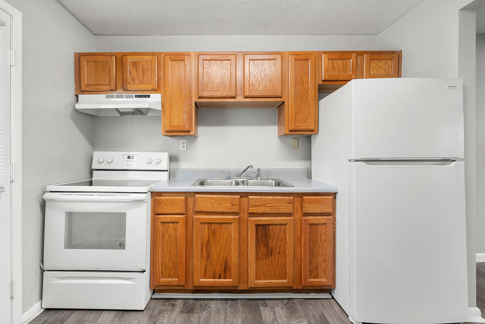 Small white kitchen with wood cabinets, white stove, sink, and refrigerator.