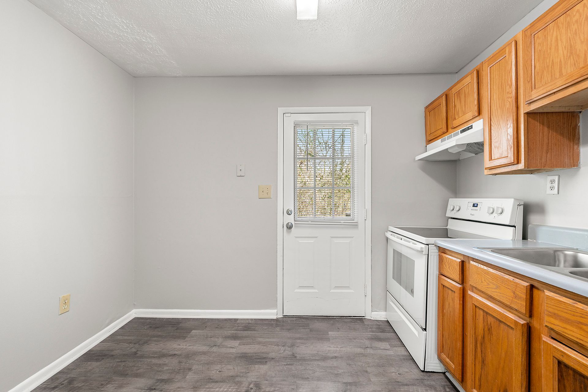 Empty kitchen with white walls, gray floor, white stove, and wooden cabinets along the right wall.