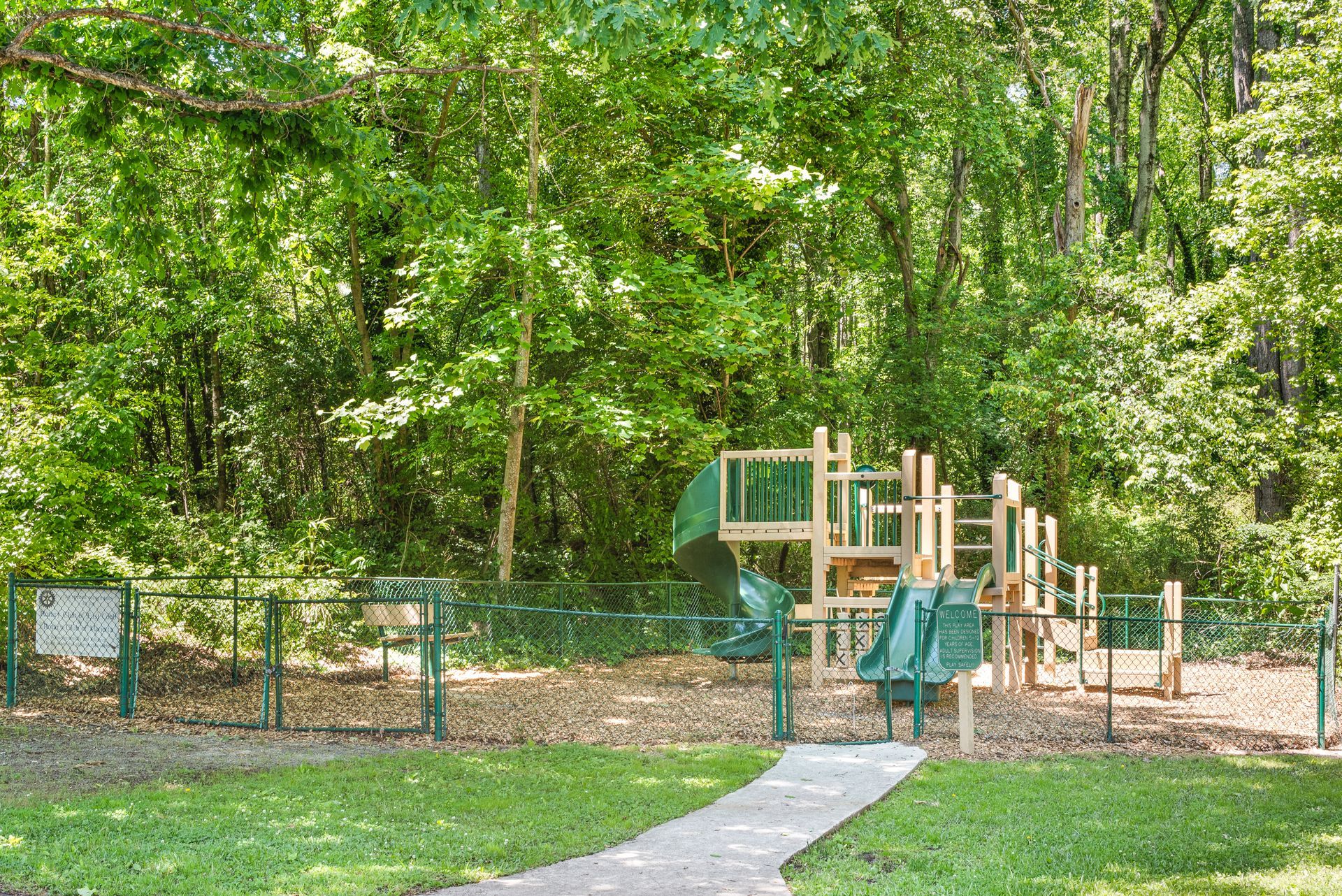 Wooded playground with green and tan climbing equipment beside a curved concrete path