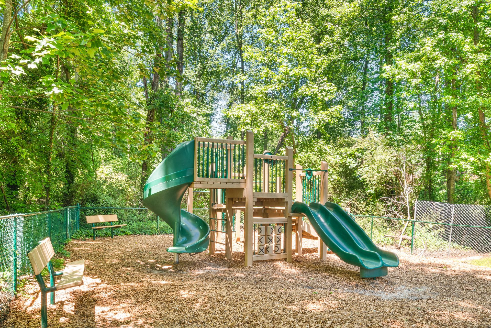 Children's playground with a green spiral slide, double slide, and wooden play structure in a wooded area