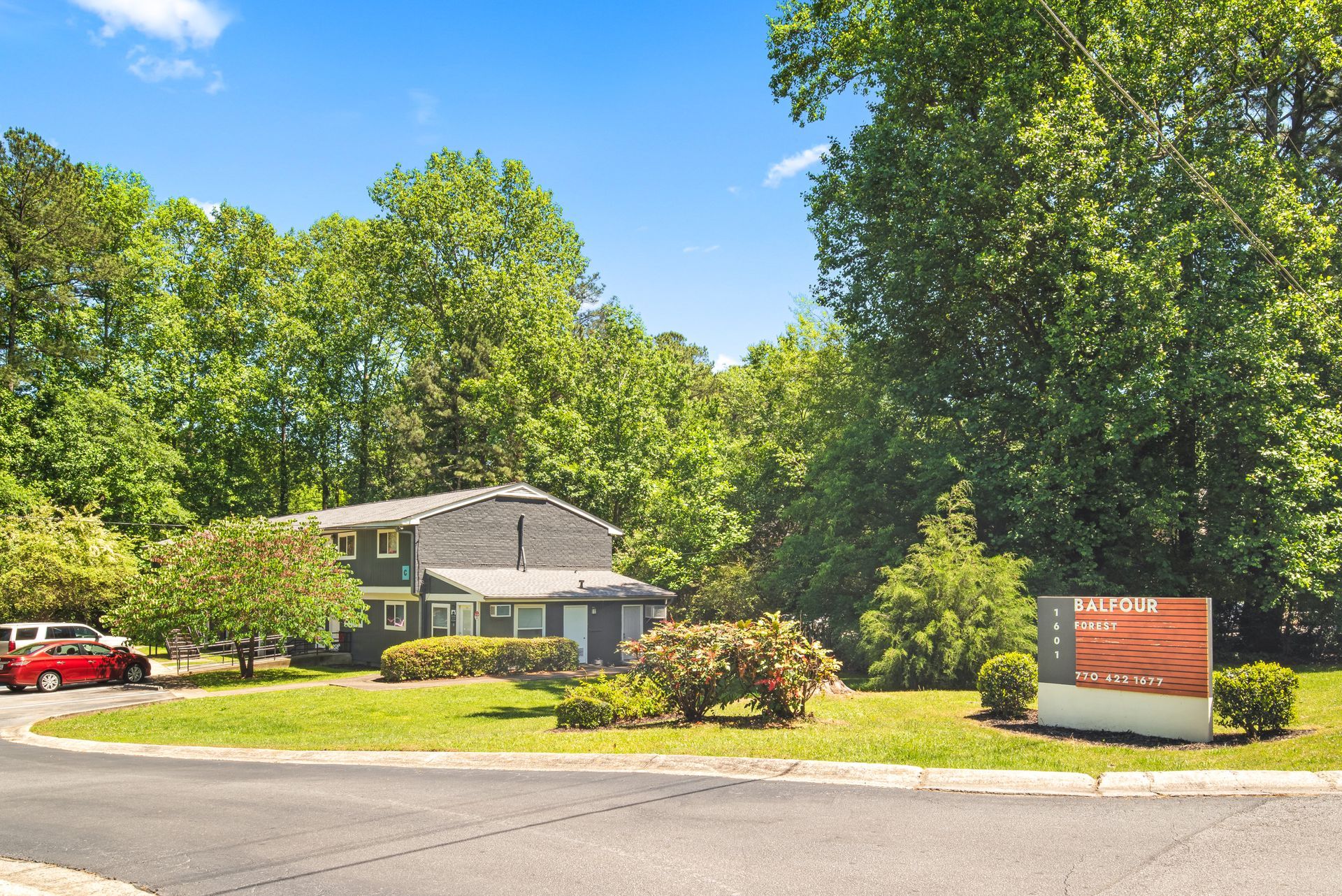 Suburban house with a manicured lawn, trees, and a red car parked nearby under a clear blue sky