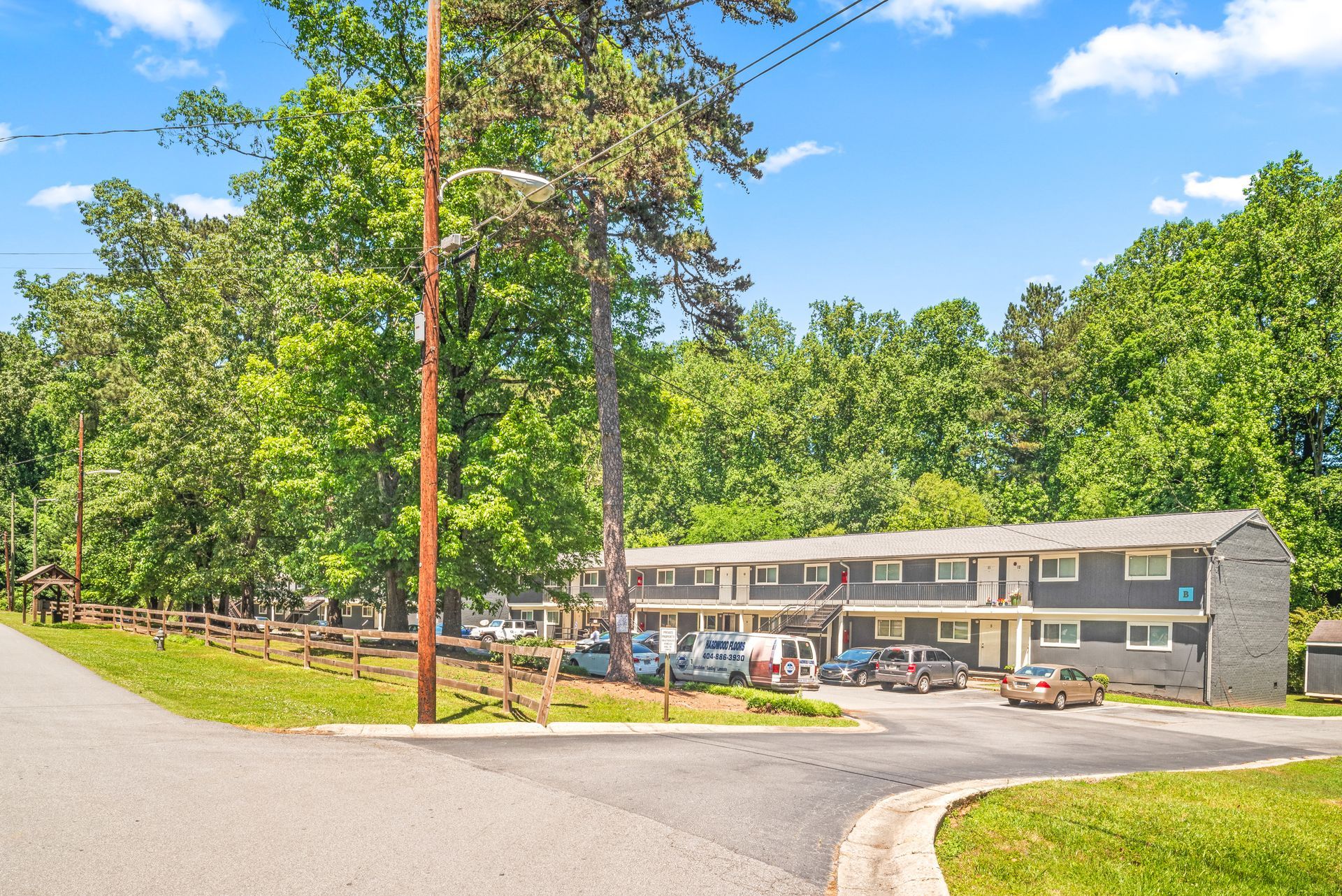 Two-story apartment building beside a curving road, with parked cars and tall trees under a blue sky