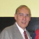Man in glasses, suit, and tie smiling, inside a room with a light-colored wall.