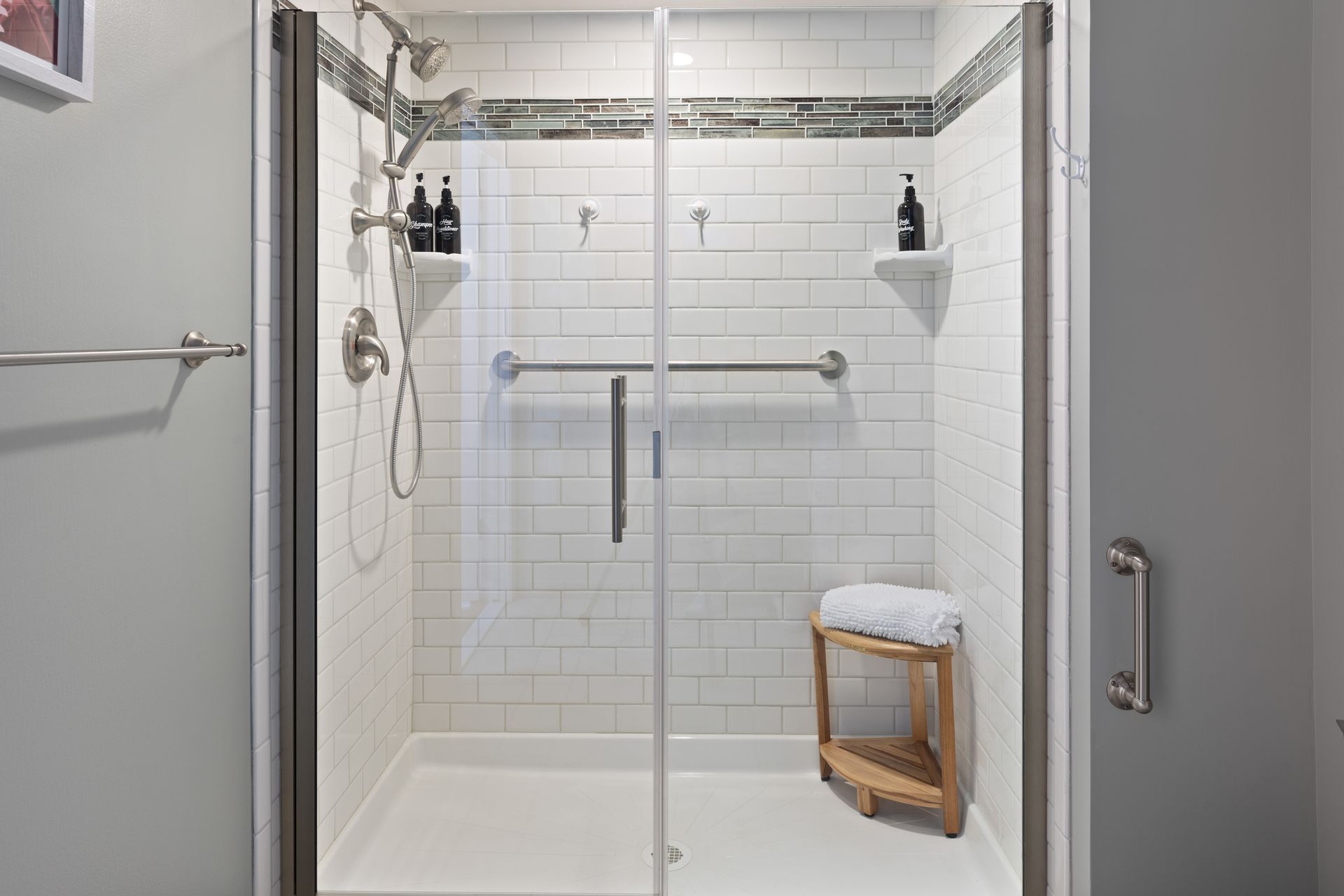 A bathroom with a walk in shower with a glass door and a wooden stool.