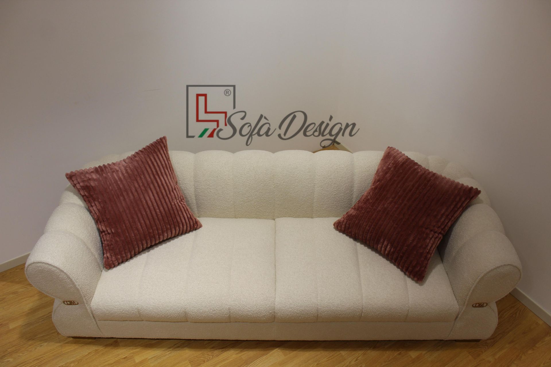 Divano color crema con due cuscini bordeaux contro una parete bianca in una stanza con pavimento in legno.