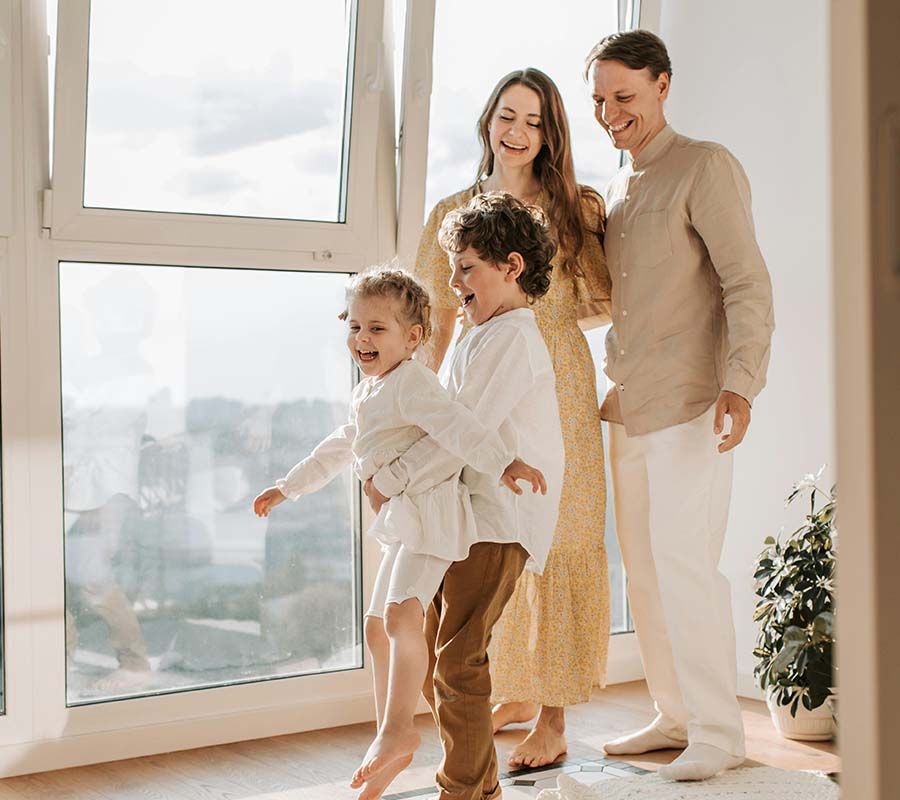 Family of four smiling by a sunlit window, with two children in the foreground and parents behind them.