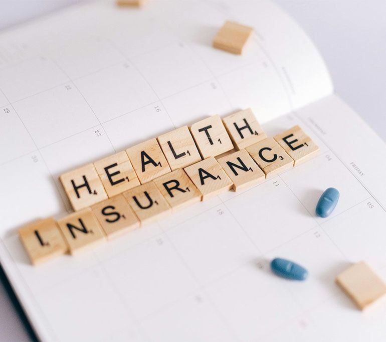 Scrabble tiles spelling “HEALTH INSURANCE” with blue capsules on a white background