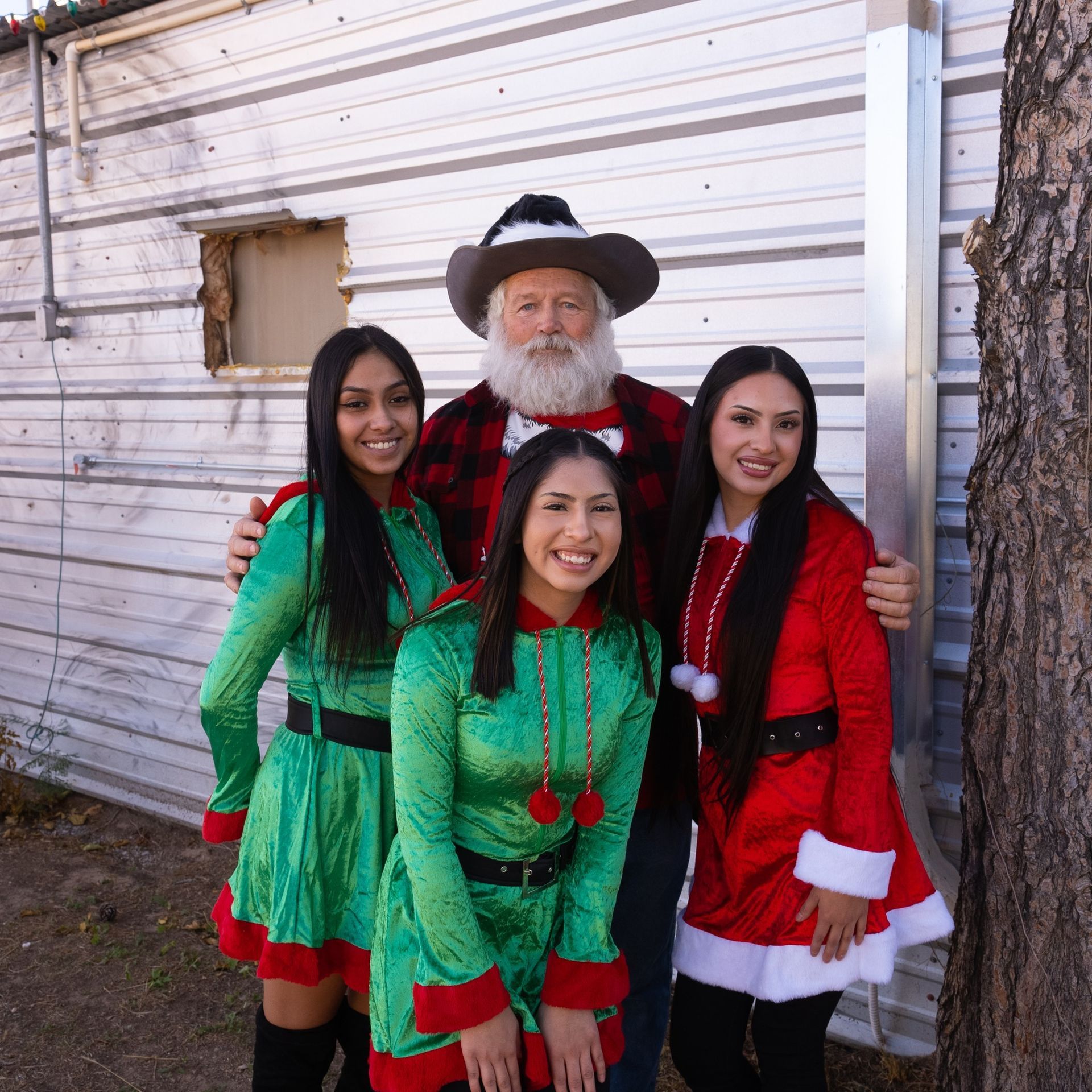 Four people in festive holiday outfits posing outside a white building beside a tree.