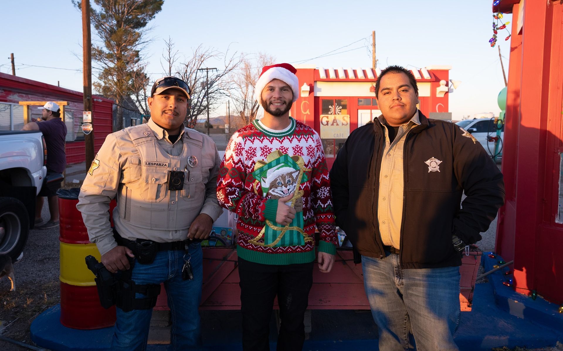 Three people standing outdoors in winter sunlight, wearing jackets and a colorful sweater, with trucks behind them.