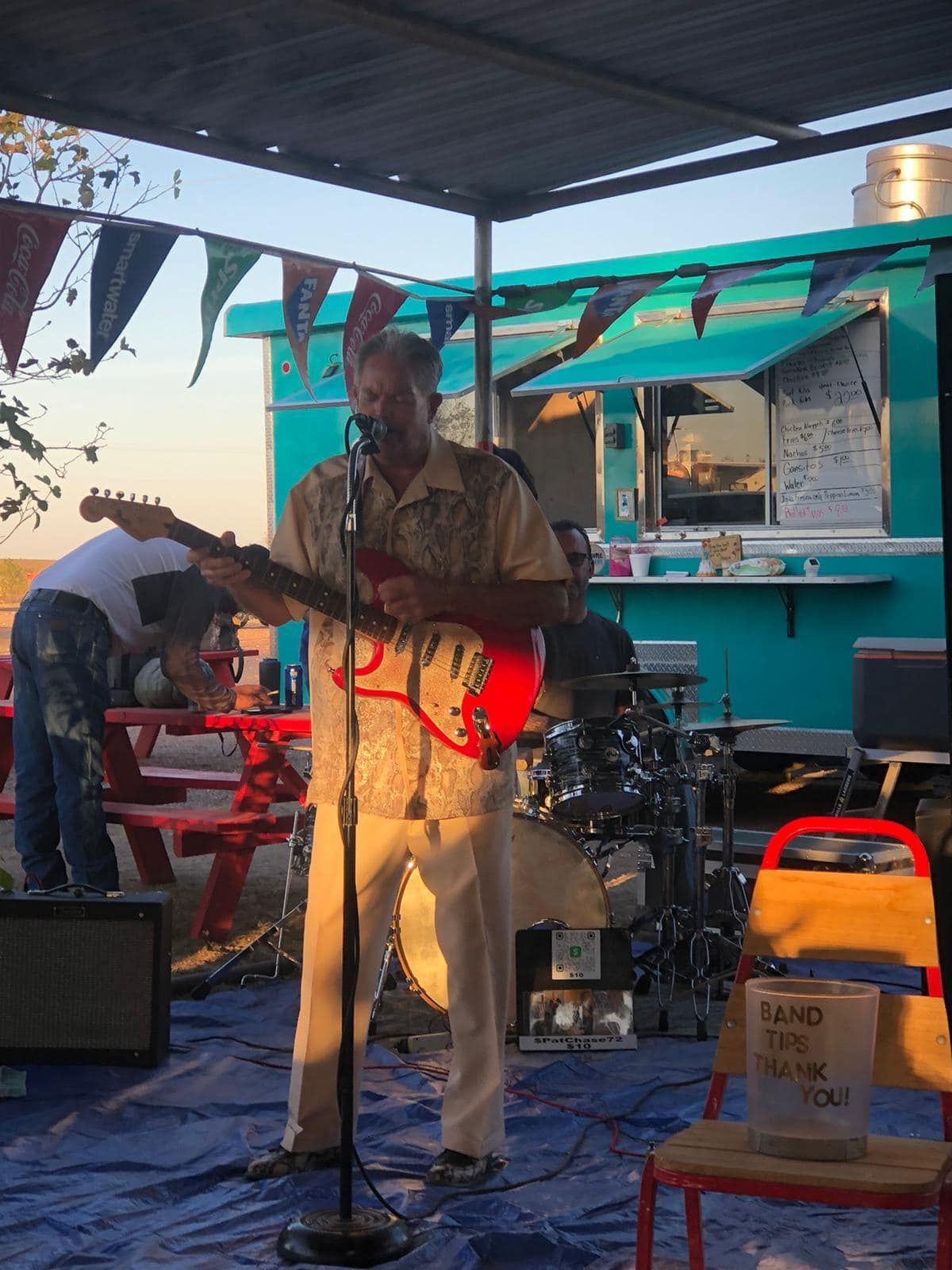 Musician singing and playing red electric guitar on a small outdoor stage under a canopy