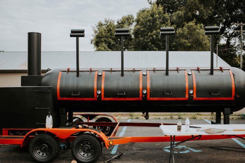 Black train-style smoker trailer with orange trim and two wheels parked outdoors.