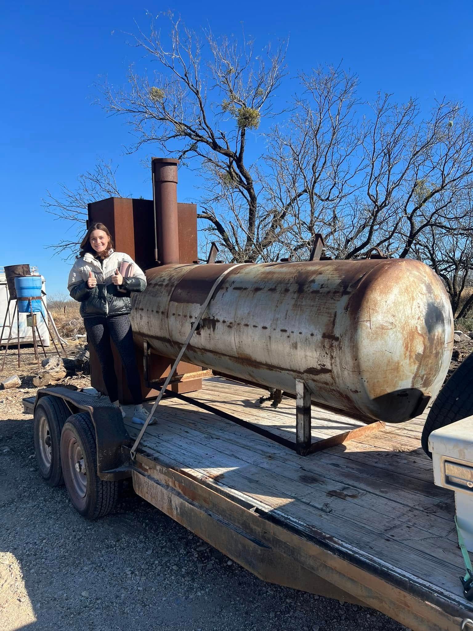 Person stands beside a rusty cylindrical tank on a trailer in a gravel yard under a clear blue sky.