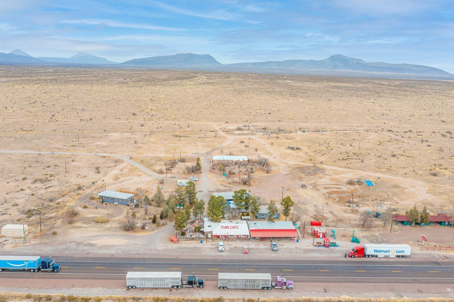 Aerial view of a roadside desert rest stop with trucks, small buildings, and sparse palm trees under a blue sky