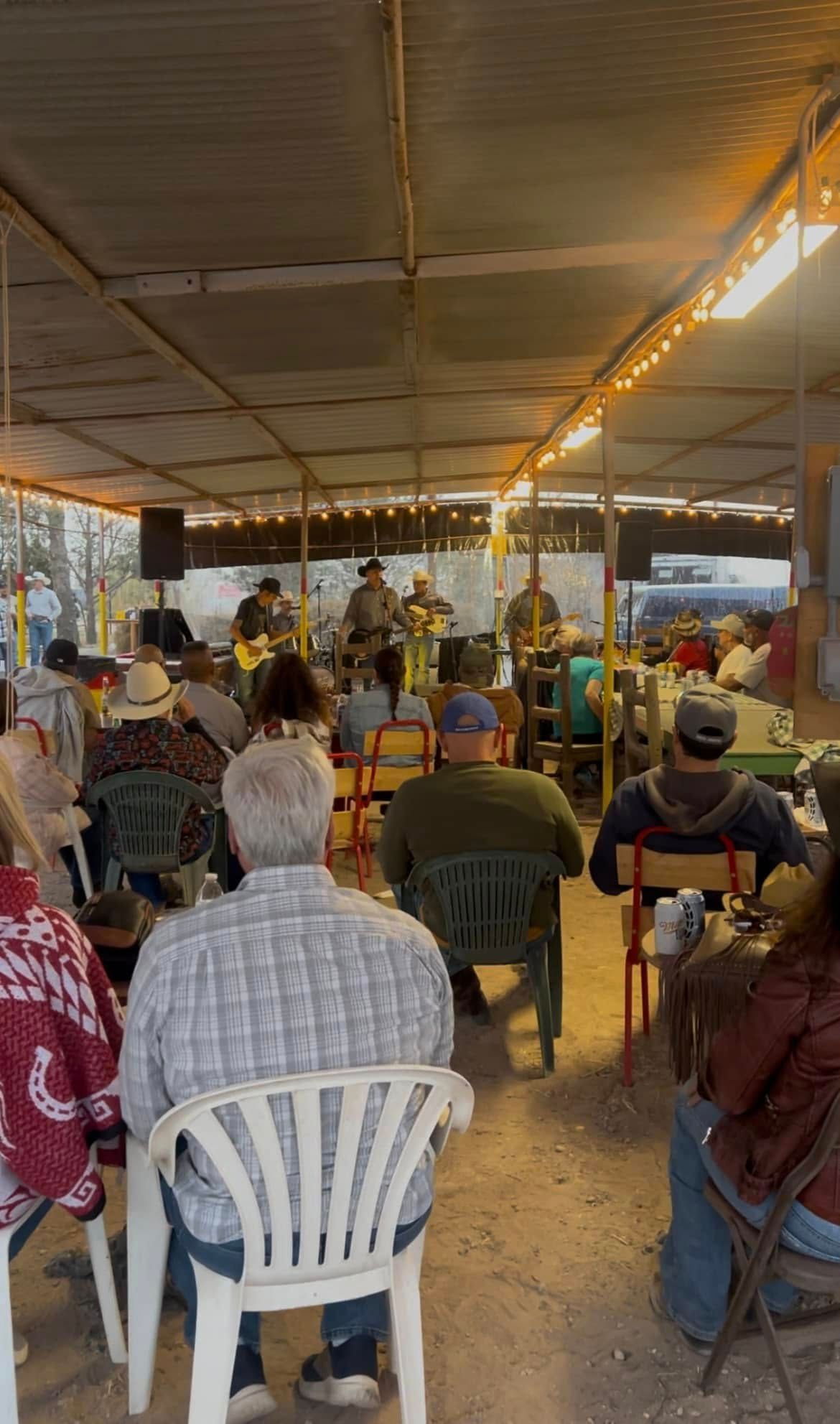 People seated under a canopy at an outdoor event, facing a speaker near the front.
