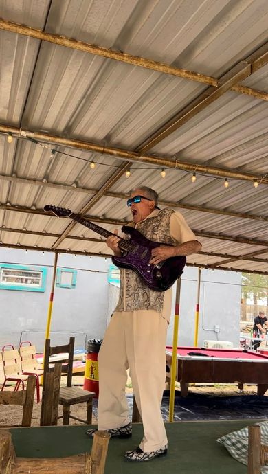 Man playing guitar on a covered outdoor stage, wearing a purple shirt and beige pants