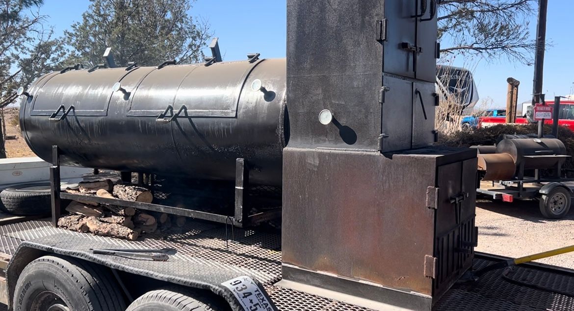Black asphalt tanker trailer parked in a lot, with a rusty brown cab and trees in the background