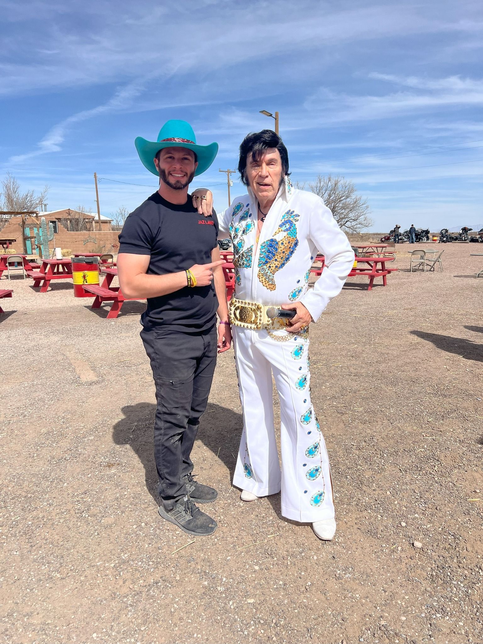Two people in a gravel lot, one in a white embroidered suit and one in a black shirt and teal hat, posing together.